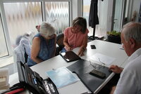 Zwei Frauen beugen sich über einen Laptop, ein älterer Mann betrachtet ein Handy.