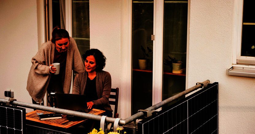 Mit PV und Köpfchen zur leistbaren Energiewende. Eine Frau sitzt auf ihrem Balkon an ihrem Laptop. Eine andere Frau steht neben ihr , hält eine Kaffeetasse in der Hand und schaut auf den Bildschirm des Laptops- An der Balkonumrandung ist eine PVC-Anlage angebracht.