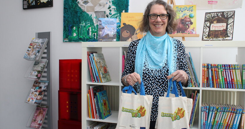 Büchereileiterin Anja Stark mit Lesestart-Sets. Büchereileiterin Anja Stark steht vor einem Bücherregal und hat Taschen mit Lesestart-Sets in der Hand.