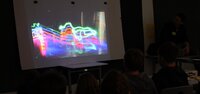 Auf einer Leinwand ist ein Foto mit bunten Lichtstreifen zu sehen. Davor sitzen Kinder und schauen sich das Bild an. In dem dunklen Raum sind nur ihre Köpfe von hinten zu erkennen..