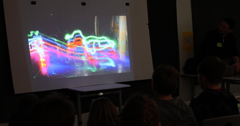 Auf einer Leinwand ist ein Foto mit bunten Lichtstreifen zu sehen. Davor sitzen Kinder und schauen sich das Bild an. In dem dunklen Raum sind nur ihre Köpfe von hinten zu erkennen..