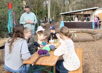 Draußen auf dem Gelände des Naturkindergartens sitzen an einem langen Tisch Kinder und Erwachsene. Auf dem Tisch liegen Stöcke, Wolle und Perlen. Ein Junge fädelt gerade Perlen auf eine Schnur.