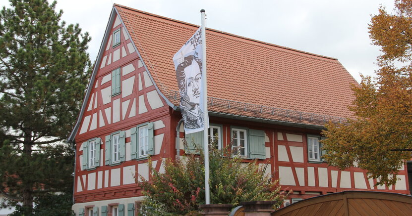 Zu sehen ist die Frontseite des Büchnerhauses, ein altes Fachwerkhaus mit roten Balken. Vor dem Haus weht eine Fahne mit dem gezeichneten Porträt Georg Büchners.