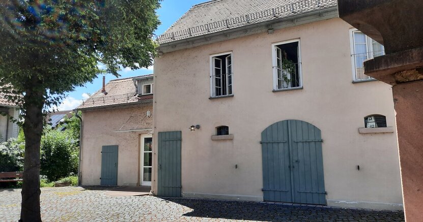 Das Kulturbüro befindet sich über der Kunstgalerie am Büchnerhaus. In dem hell verputzten Haus befindet sich im Erdgeschoss mit den blauen Holzläden die Kunstgalerie und im ersten Stock das Kulturbüro. Dort ist das mittlere von drei Fenstern geöffnet. Links im Vordergrund steht ein Baum.