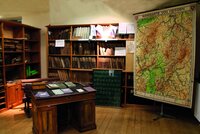 Zu sehen ist ein altes Klassenzimmer: Im Vodergrund steht ein altes Pult mit alten Heften und Büchern. An der Wand befinden sich alte Holzschränke mit Büchern. Rechts im Hintergrund hängt eine alte Wandkarte von Hessen.