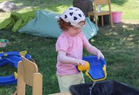 Mit Wasser planschen stand angesichts der Temperaturen hoch im Kurs. Ein kleines Mädchen mit rosa T-Shirt und weißem Sonnenkäppi steht vor einem Trog, der mit Wasser gefüllt ist. Sie hat in der einen Hand eine bkaze Schale, in die sie mit der andernen Hand aus einem gelben Becher Wasser gießt.