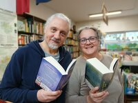 Oliver Bopp und Claudia Borek von der Buchhandlung Faktotum stehen in ihrer Buchhandlung. Sie halten jeweils ein aufgeschlagenes Buch in der Hand und lächeln in die Kamera.