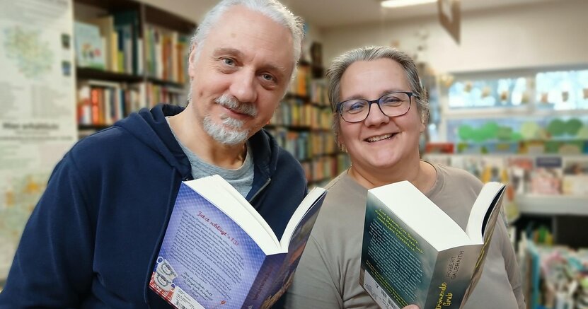 Oliver Bopp und Claudia Borek von der Buchhandlung Faktotum stehen in ihrer Buchhandlung. Sie halten jeweils ein aufgeschlagenes Buch in der Hand und lächeln in die Kamera.