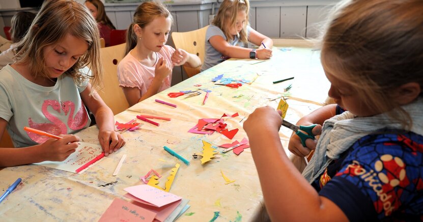 Kinder sitzen an beiden Seiten eines langen Tisches und malen und schneiden aus.