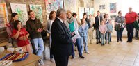 Erster Stadtrat Richard Kraft steht im Anzug in der Kunstgalerie am Büchnerhaus. An der Wand sind bunte Kindergemälde zu sehen. Davor stehen Eltern mit ihren Kindern und hören Kraft zu. An der Seite steht ein Tisch mit Kleinigkeiten zum Essen.
