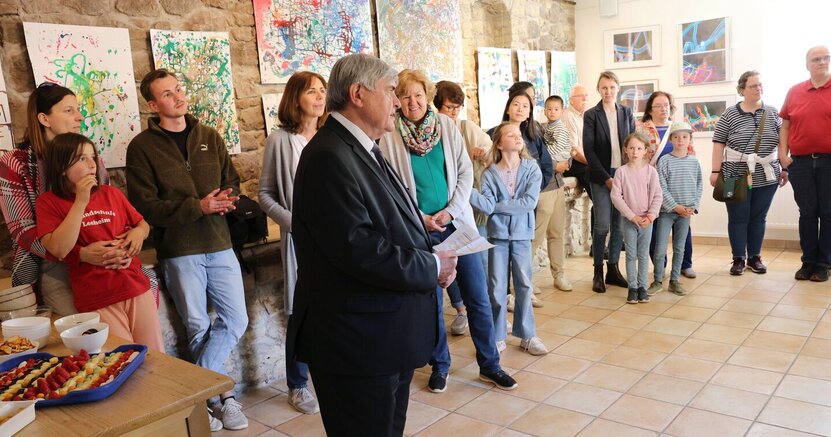 Erster Stadtrat Richard Kraft steht im Anzug in der Kunstgalerie am Büchnerhaus. An der Wand sind bunte Kindergemälde zu sehen. Davor stehen Eltern mit ihren Kindern und hören Kraft zu. An der Seite steht ein Tisch mit Kleinigkeiten zum Essen.