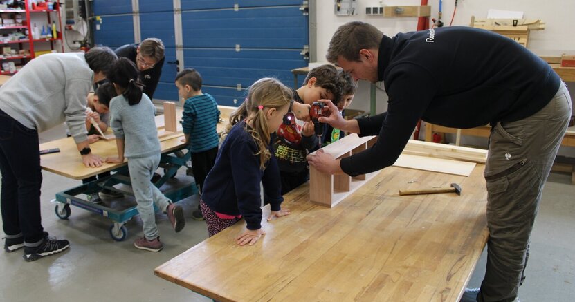 In der Schreinerei stehen ein Mädchen und zwei Jungs auf einer Seite einer Arbeitsplatte. Auf der anderen Seite steht André Lindemann. Er hilft dem Jungen in der Mitte, mit einem Akkubohrer eine Schraube in einen Holzkasten zu schrauben.