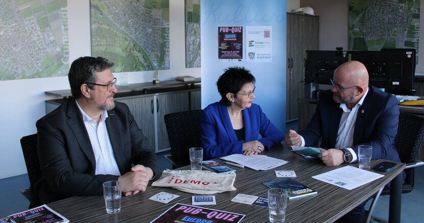 Bürgermeister Marcus Kretschmann (rechts) im Gespräch mit Kulturbüroleiter Marco Hardy und Ute Wiegand-Fleischhacker, Landes- und Kreisvorsitzende der Europa-Union. Bürgermeister Marcus Kretschmann (rechts) sitzt mit Kulturbüroleiter Marco Hardy und Ute Wiegand-Fleischhacker, Landes- und Kreisvorsitzende der Europa-Union an einem Tisch und redet. Im Hintergrund ist ein Plakat vom Pub Quiz zu sehen.