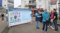 Mitten in Riedstadt möchte die Ausstellung die Stimmen von Menschen mit Behinderung sichtbar machen. Auf dem Rathausplatz steht ein großes Banner. Auf dem steht "Menschenrechte sind nicht verhandelbar". Darunter sind viele bunte Handabdrücke zu sehen. Sie bilden zusammen einen Kreis. Vor dem Banner stehen Menschen und unterhalten sich.