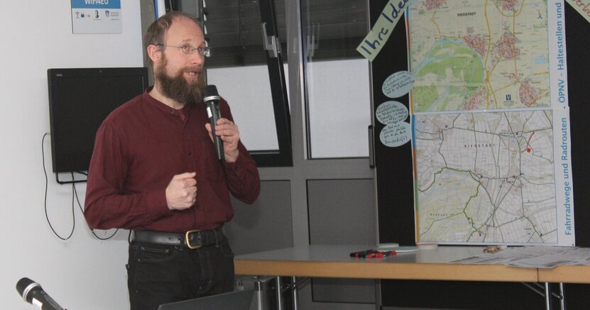 Sanierungsmanager Benjamin Krick freut sich auf Verstärkung im Sanierungsmanagement der Stadt. Sanierungsmanager Benjamin Krick steht bei einer Veranstaltung im Sitzungssaal des Riedstädter Rathaus mit Mikrofon in der Hand neben einer Stellwand mit zwei Karten. Links und rechts der oberen Karte sind zwei Zettel befestigt. Auf dem einen steht "Ihre Ideen" und auf dem anderen "Rad- und Nahverkehr".