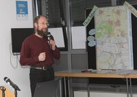 Sanierungsmanager Benjamin Krick freut sich auf Verstärkung im Sanierungsmanagement der Stadt. Sanierungsmanager Benjamin Krick steht bei einer Veranstaltung im Sitzungssaal des Riedstädter Rathaus mit Mikrofon in der Hand neben einer Stellwand mit zwei Karten. Links und rechts der oberen Karte sind zwei Zettel befestigt. Auf dem einen steht "Ihre Ideen" und auf dem anderen "Rad- und Nahverkehr".