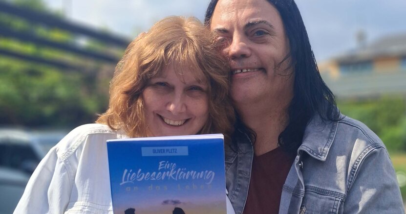 Steffi und Oliver Pletz mit dem Buch „Eine Liebeserklärung an das Leben“ Steffi und Oliver Pletz stehen draußen auf einem Platz. Im Hintergrund ist Grün und der Himmel zu sehen. Sie halten das Buch "Eine Liebeserklärung an das Leben" in den Händen.