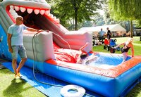 Eine Riesen-Wasserrutsche war die Attraktion für die Kinder. Auf der Liegewiese steht eine aufblasbare Wasserrutsche in Form eines Fischkopfes mit Zähnen. Ein Mann spritzt mit einem Schlauch Wasser auf die Rutsche. Ein Junge landet gerade unten im Wasser, ein Mädchen klettert gerade aus dem Becken.
