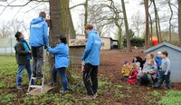 Ein Mann mit blauer Jacke und der weißen Aufschrift "Nabu" steht auf einer Leiter und bringt an einem Baum einen Nistkasten an. Um ihn herum stehen eine Frau und zwei Männer vom Nabu. Kitaleiterin Anja Llorca und Viktoria Orban-Radics kauern mit einigen Kindern am Boden und beobachten die Aktion.