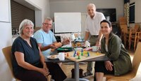 Spielend gegen die Einsamkeit: Karin Schnatbaum, Ottmar Eberling, Dr. Eckhardt Stein und Stefanie Drozdzynski (v.li.) im Jugendraum der SKG Erfelden, dem Ort des Spieletreffs. Karin Schnatbaum, Ottmar Eberling und Stefanie Drozdzynski sitzen an einem Tisch im Jugendraum der SKG Erfelden. Dr. Eckhardt Stein steht am Tisch. Auf dem Tisch ist ein Mensch-ärgere-dich-nicht Spiel aufgebaut. Ottmar Eberling hält einen Würfelbecher in der Hand. Auf einem weiteren Tisch im Hintergrund sind eine Kaffeekanne, Tassen, Wasserflaschen und Gebäck zu sehen.