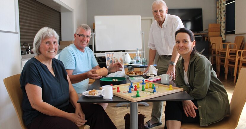 Spielend gegen die Einsamkeit: Karin Schnatbaum, Ottmar Eberling, Dr. Eckhardt Stein und Stefanie Drozdzynski (v.li.) im Jugendraum der SKG Erfelden, dem Ort des Spieletreffs. Karin Schnatbaum, Ottmar Eberling und Stefanie Drozdzynski sitzen an einem Tisch im Jugendraum der SKG Erfelden. Dr. Eckhardt Stein steht am Tisch. Auf dem Tisch ist ein Mensch-ärgere-dich-nicht Spiel aufgebaut. Ottmar Eberling hält einen Würfelbecher in der Hand. Auf einem weiteren Tisch im Hintergrund sind eine Kaffeekanne, Tassen, Wasserflaschen und Gebäck zu sehen.