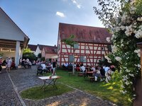 Treffen, Genießen und Bleiben: Begegnungen beim Woi-Eck am BüchnerHaus Tische und Bänke stehen auf dem gepflasterten Hof und dem Rasen vor einem alten Fachwerkhaus - dem Büchnerhaus. An den Tischen sitzen viele Menschen und unterhalten sich. Im Bildvordergrund ist ein weiß blühender Busch zu sehen.