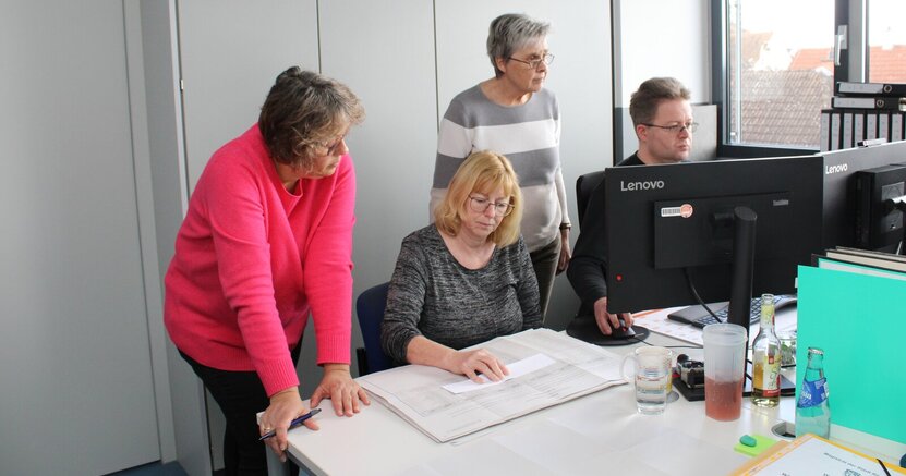 Cornelia Anthes sitzt in einem Raum des Rathauses am Tisch und hat einen Stimmzettel vor sich liegen. Alexandra Hartmann steht neben ihr und schaut auch auf den Zettel. Daneben sitzt Peter Kraus und gibt Zahlen in den PC en, Bärbel Fischer steht hinter ihm und schaut, was er eingibt.