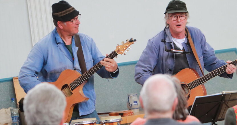 Franz Offenbecher und Klaus (v.li.) stehen mit ihren Gitarren vor ihrem Publikum.  Von den Zuhörerinnen und Zuhörern sind einige Hinterköpfe zu sehen.
