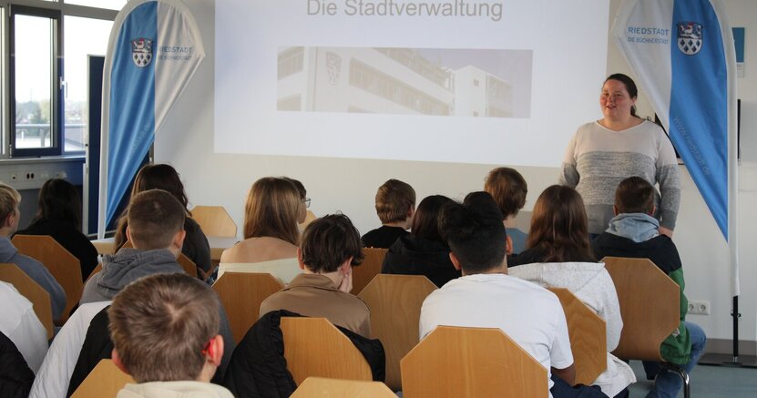 Ausbildungsleiterin Julia Galley gab einen Überblick über die Aufgaben einer Stadtverwaltung Jungen und Mädchen sitzen in Stuhlreihen mit dem Rücken zum Betrachter im Rathaussaal. Vor ihnen steht Ausbildungsleiterin Julia Galley. Neben ihr ist an der Wand eine Seite der Präsentation mit der Schlagzeile "Die Stadtverwaltung" zu sehen.