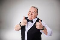 Woody Feldmann kommt nach Riedstadt. Woody Feldmann hat ein weißes Hemd mit schwarzer Fliege und eine schwarze Weste an. Sie streckt beide Daumen nach oben.