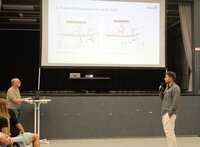 Paul Javier Schüle vom Planungsbüro Sweco gab Informationen zu den Änderungen für den zweiten Ausschreibungsabschnitt. Paul Javier Schüle vom Planungsbüro Sweco steht mit Mikrofon vor der Bühne der Heinrich-Bonn-Halle. Auf der Bühne ist eine Leinwand aufgebaut. Er gibt Erläuterungen zu den Bildern, die auf der Leinwand zu sehen sind.