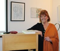 Suzanne Bohn bei ihrem Vortrag im letzten Jahr. Suzanne Bohn lehnt sich gegen ein Stehpult, im Hintergrund sind gerahmte Bilder zu sehen. Das Foto entstand letztes Jahr in der Kunstgalerie am Büchnerhaus.