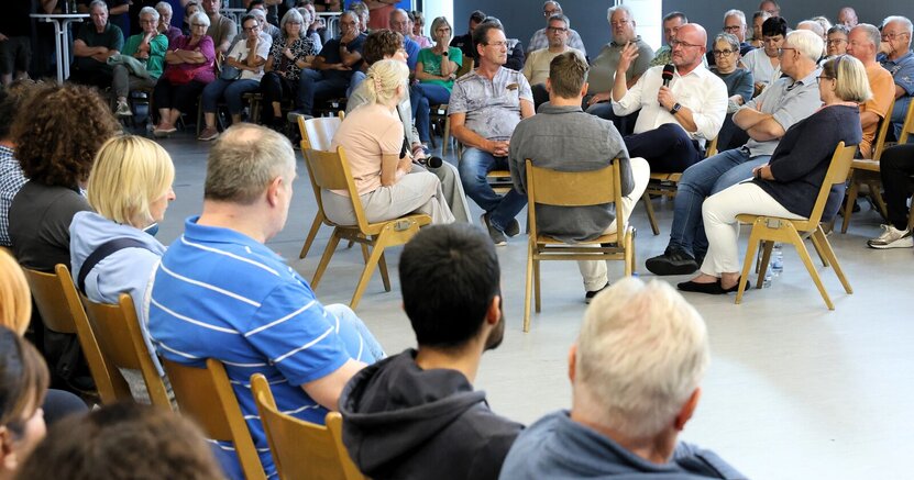 In der Gesprächsrunde konnten auch immer zwei Menschen mit Fragen oder Anliegen Platz nehmen. Mit Mikrofon Bürgermeister Marcus Kretschmann, rechts von ihm Fachbereichsleiter Bauen Joachim Götz und Stadtwerkeleiterin Saskia Kirsch. In der Heinrich-Bonn-Halle sitzen in einem Stuhlkreis Bürgermeister Marcus Kretschmann und weitere Personen. Drum herum sitzen auf Stühlen viele Zuhörer, einige stehen auch an Stehtischen.