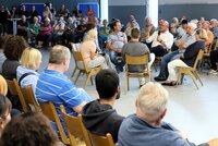 In der Gesprächsrunde konnten auch immer zwei Menschen mit Fragen oder Anliegen Platz nehmen. Mit Mikrofon Bürgermeister Marcus Kretschmann, rechts von ihm Fachbereichsleiter Bauen Joachim Götz und Stadtwerkeleiterin Saskia Kirsch. In der Heinrich-Bonn-Halle sitzen in einem Stuhlkreis Bürgermeister Marcus Kretschmann und weitere Personen. Drum herum sitzen auf Stühlen viele Zuhörer, einige stehen auch an Stehtischen.