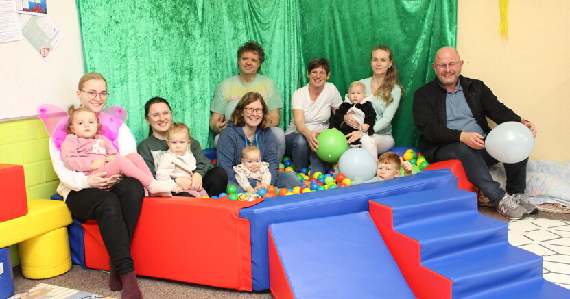 In einem Bällebad mit bunten Bällen sitzen und liegen kleine Kinder. Drumherum sitzen auf der Umrandung Mütter mit ihren Kindern, die Gruppenbetreuer Heike Schuhknecht, Mario Quast und Katharina Jantzen sowie Bürgermeister Marcus Kretschmann.