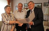 Helga Schneider gibt lächelnd dem Ersten Stadtrat Richard Kraft die Hand. Dieser hält eine Mappe mit Glückwünsch-Urkunden in der Hand. Zwischen ihnen steht Wilhelm Schneider.