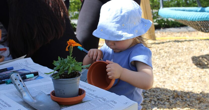 Ein kleines Mädchen mit Sonnenhut bemalt mit einem Filzstift einen Blumentopf. Vor ihr stehen auf einem Tisch eine Pflanze mit oranger Blüte und eine Gartenschaufel.