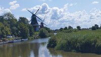 Die Greetsieler Zwillingsmühlen gehören auch zum Besichtigungsprogramm In der Bildmitte ist ein Fluss zu sehen. Links und rechts an seinem Ufer stehen Büsche und Bäume. Links stehen hintereinander zwei gleich aussehende Windmühlen.