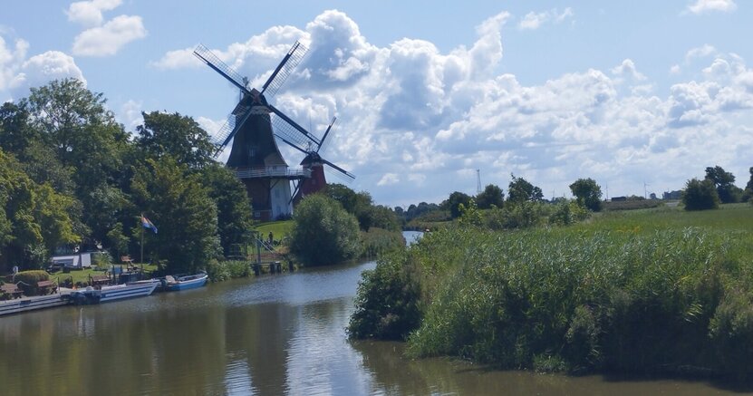In der Bildmitte ist ein Fluss zu sehen. Links und rechts an seinem Ufer stehen Büsche und Bäume. Links stehen hintereinander zwei gleich aussehende Windmühlen.