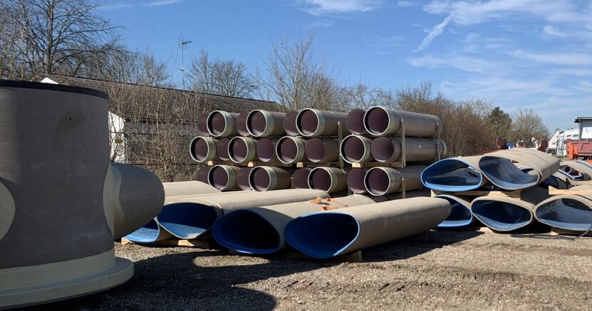 Auf einem Lagerplatz liegen mehrere Kanalrohre auf dem Boden. Sie sind nicht rund, sondern oval und laufen unten spitz zu. Innen sind sie blau ausgekleidet. Im Hintergrund sind normale runde Kanalrohre übereinander gestapelt.