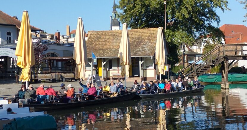 Mehrere lange Kanus haben längs an einer Uferpromenade festgemacht. In den Booten sitzen viele Menschen.
