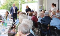 An festlich gedeckten Tischen sitzen ältere Menschen. Vor ihnen steht Bürgermeister Marcus Kretschmann mit Mikrofon und spricht. Neben ihm steht die Leiterin der Seniorenberatungsstelle Stefanie Drozdzynski.
