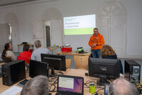 Von hinten sind Menschen zu sehen, die an Tischen vor ihren aufgeklappten Laptops sitzen. Vorne steht ein Mann und erklärt etwas. An der Wand steht "Willkommen zum 2. Linux-Café"