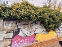 Ein Beispiel aus dem traditionellen Auszeit-Berlin-Fotowettbewerb Zu sehen ist eine bemalte Mauer in Berlin. Darauf sind ein Männer- und ein Frauenkopf abgebildet. Die Büsche auf der Mauer sehen aus wie Haare.