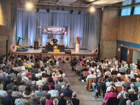Auf der Bühne der Christoph-Bär-Halle ist Woody Feldmann unter einem großen Banner mit ihrem Namen zu sehen. Links ist aufblasbares Schwimmzubehör wie ein Krokodil und ein Schwimmring zu sehen. Rechts große Badenudeln. In der Halle sitzen viele Menschen in den vollbesetzten Reihen. 