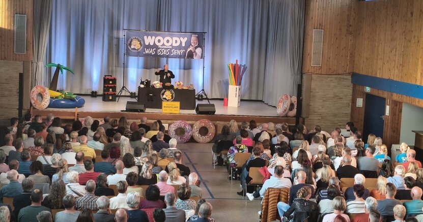 Woody Feldmann sorgte für eine volle Christoph-Bär-Halle Auf der Bühne der Christoph-Bär-Halle ist Woody Feldmann unter einem großen Banner mit ihrem Namen zu sehen. Links ist aufblasbares Schwimmzubehör wie ein Krokodil und ein Schwimmring zu sehen. Rechts große Badenudeln. In der Halle sitzen viele Menschen in den vollbesetzten Reihen.