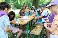 Aus Wollfäden werden Bänder geknüpft. Einige Kinder haben Wopllfäden an einem Tisch befestigt und flechten sie zu Bändern. Andere malen mit Stiften auf weiße Stoffstreifen.