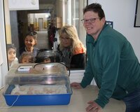 Der Brutkasten ist da. Erzieherin Felicia Burkhardt, Kitaleiterin Yvonne Dickler und Michaela Schwäch vom GZV (v.li.) staunen mit den Kindern Isaak, Zamor und Amber. Im Vordergrund steht der Brutkasten mit einer blauen Schale auf einem Tisch. In dem Brutkasten liegen verschieden große Eier. Daneben steht Michaela Schwäch und stützt sich auf dem Tisch ab. Durch eine Glasscheibe sieht man drei Kinder und zwei Erzieherinnen, die sich den Brutkasten anschauen.