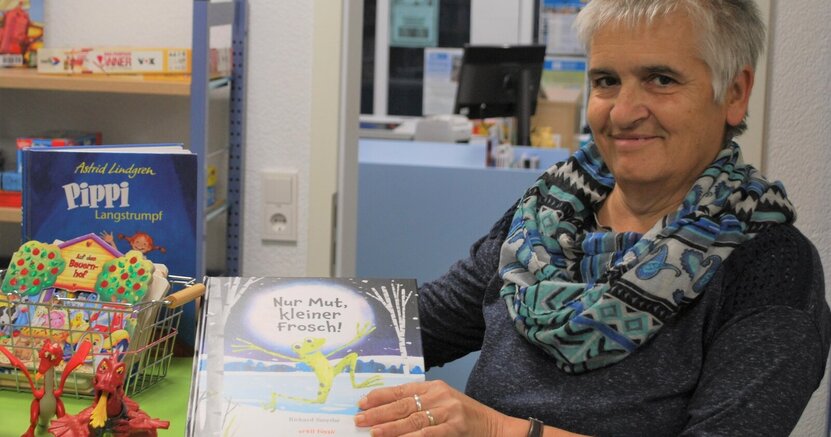 Büchereimitarbeiterin Ellen Stieb steht in der Bücherei Wolfskehlen an einem Tisch und zeigt das Bilderbuch "Nur Mut, kleiner Frosch. Auf dem Tisch stehen Tonie-Figuren in Form von Drachen. In einem Drahtkorb befinden sich weitere Kinderbücher.