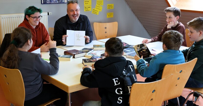 Autorin Alina Hesse und Jugendpfleger Kai Faßnacht sitzen mit Jugendlichen um einen Tisch. In der Mitte liegen viele Bücher.